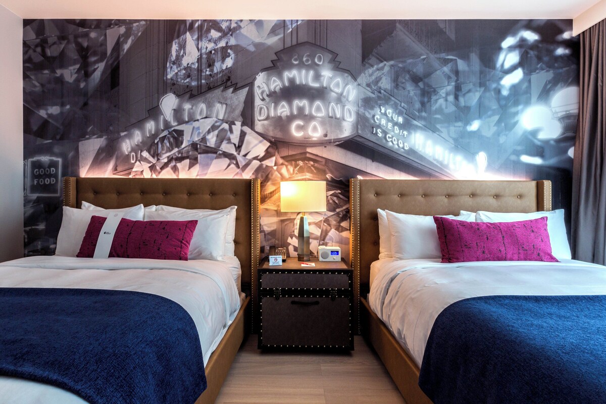 Una habitación de hotel con dos camas, una mesita de noche con lámpara y un mural en la pared.