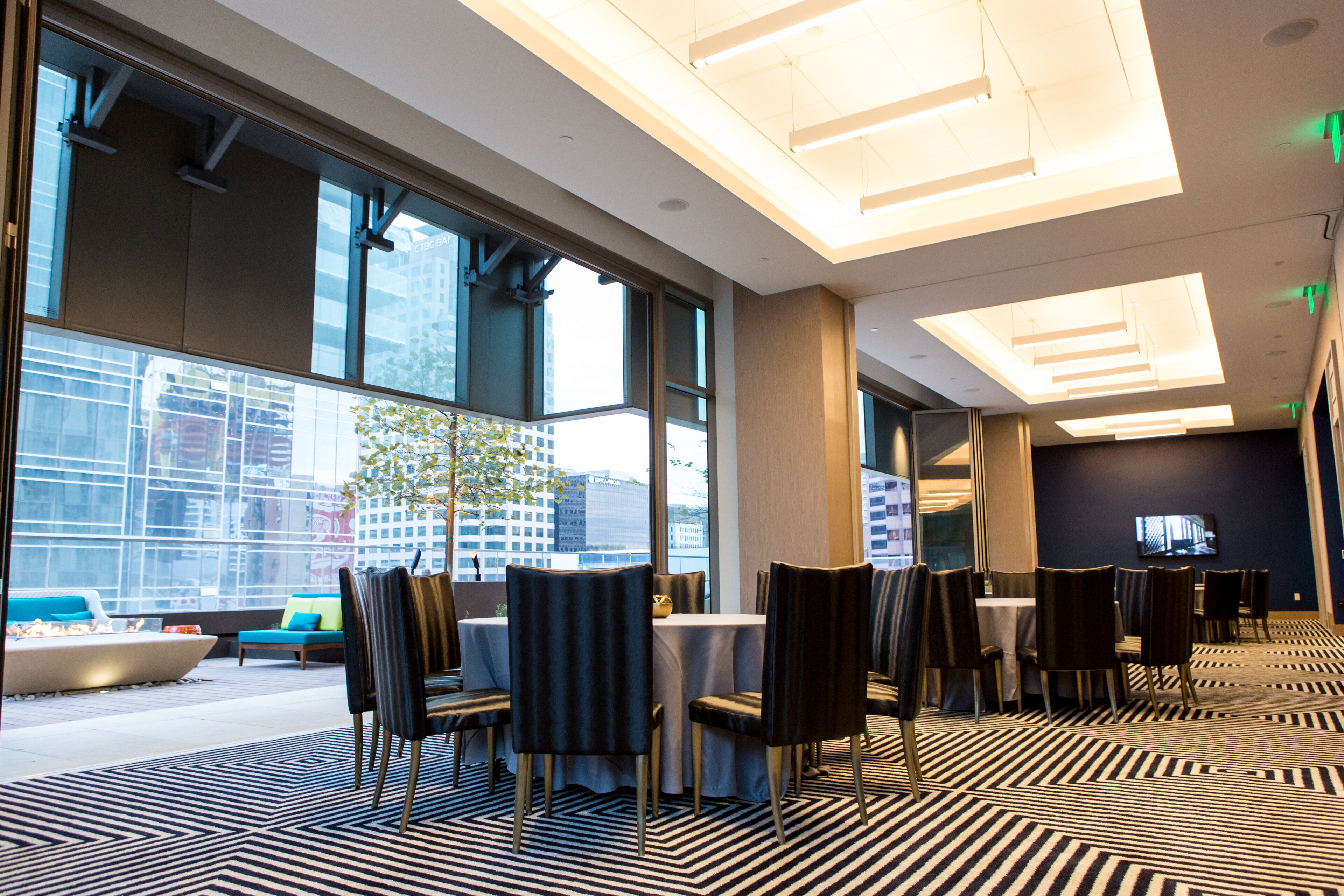 A modern hotel lobby with large windows, a patterned carpet, and a seating area with chairs and tables.
