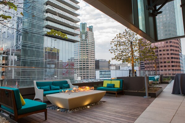 Un área de estar al aire libre con mobiliario moderno, una chimenea y vistas de edificios altos circundantes.