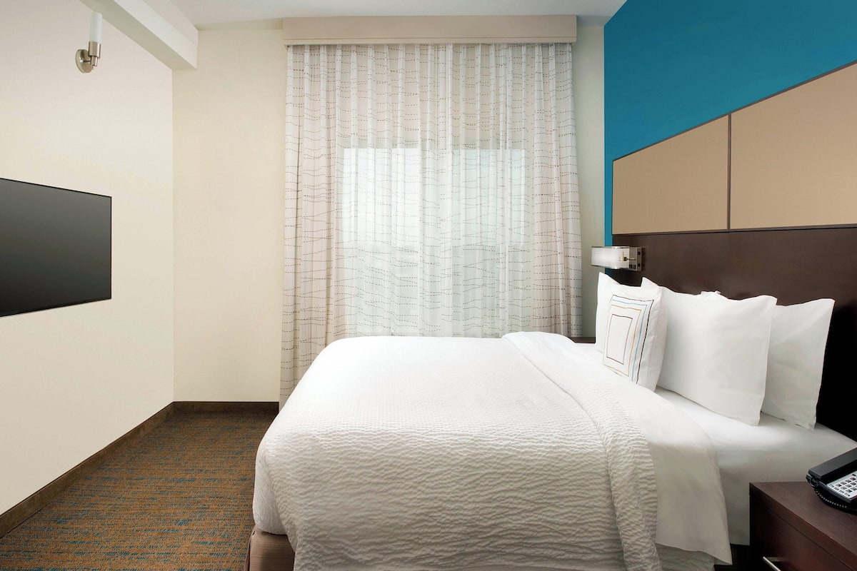 Ein Hotelzimmer mit einem großen Bett, einem Flachbildfernseher, einem Nachttisch mit Telefon und einem Fenster mit Vorhängen.