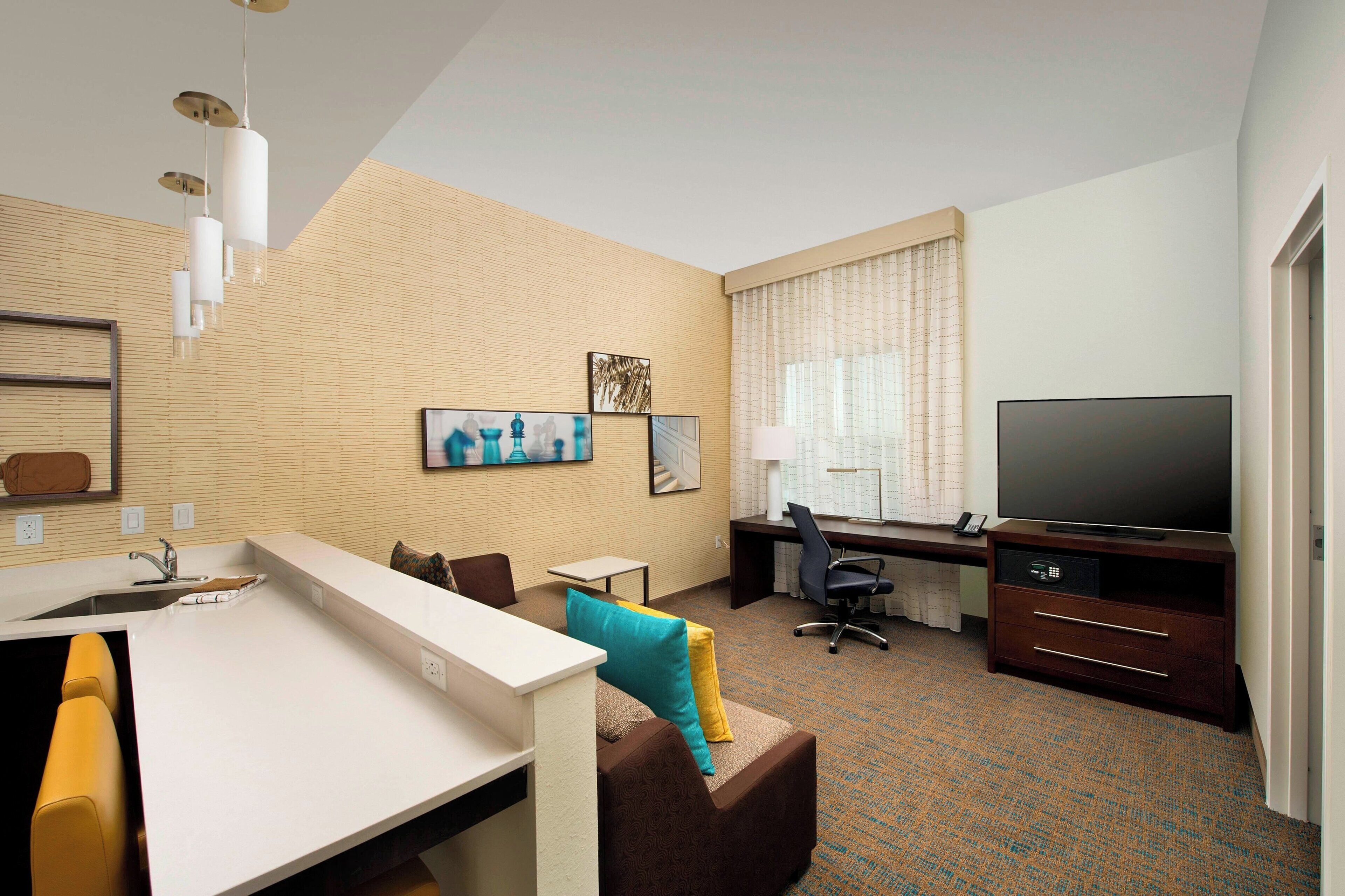 A hotel room with a kitchenette, a sofa with cushions, a desk with a chair, a television, and artwork on the wall.