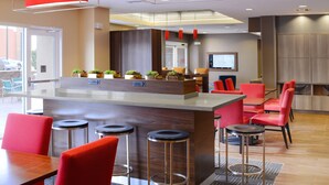 Eine moderne Hotellobby mit Barbereich in der Mitte, roten Sitzgelegenheiten und einem Bildschirm an der Wand.