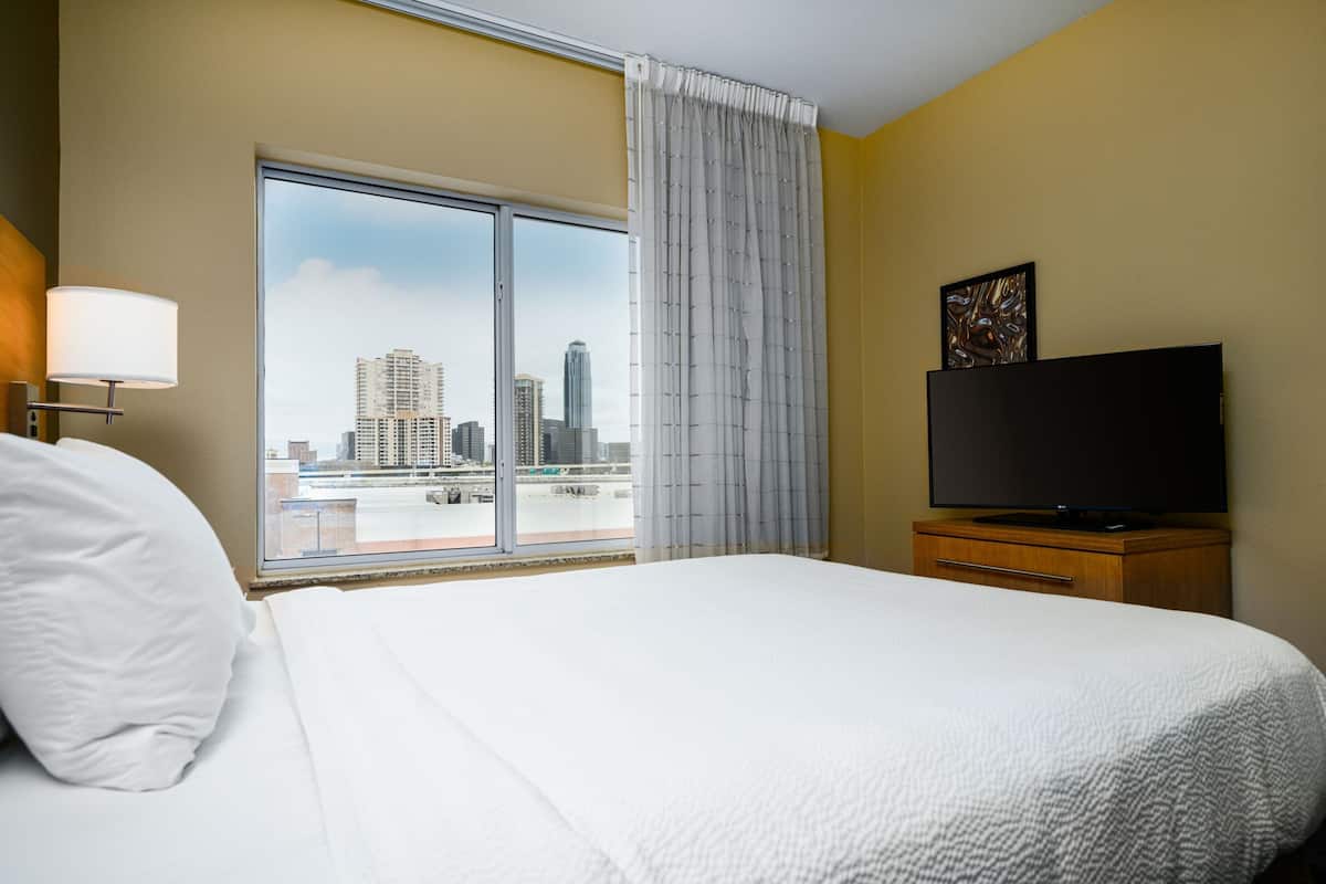 Habitación de hotel con una cama grande, un televisor de pantalla plana, un ventanal con vistas a la ciudad y una mesita de noche de madera.