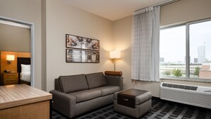 Ein Hotelzimmer mit einer Couch, einem kleinen Tisch, einer Lampe, einem Spiegel und einem Fenster mit Blick auf die Stadt.