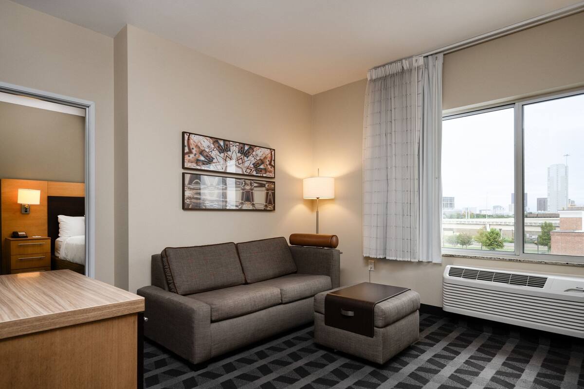 Habitación de hotel con sofá, una mesita, lámpara, espejo y ventana con vista a la ciudad.