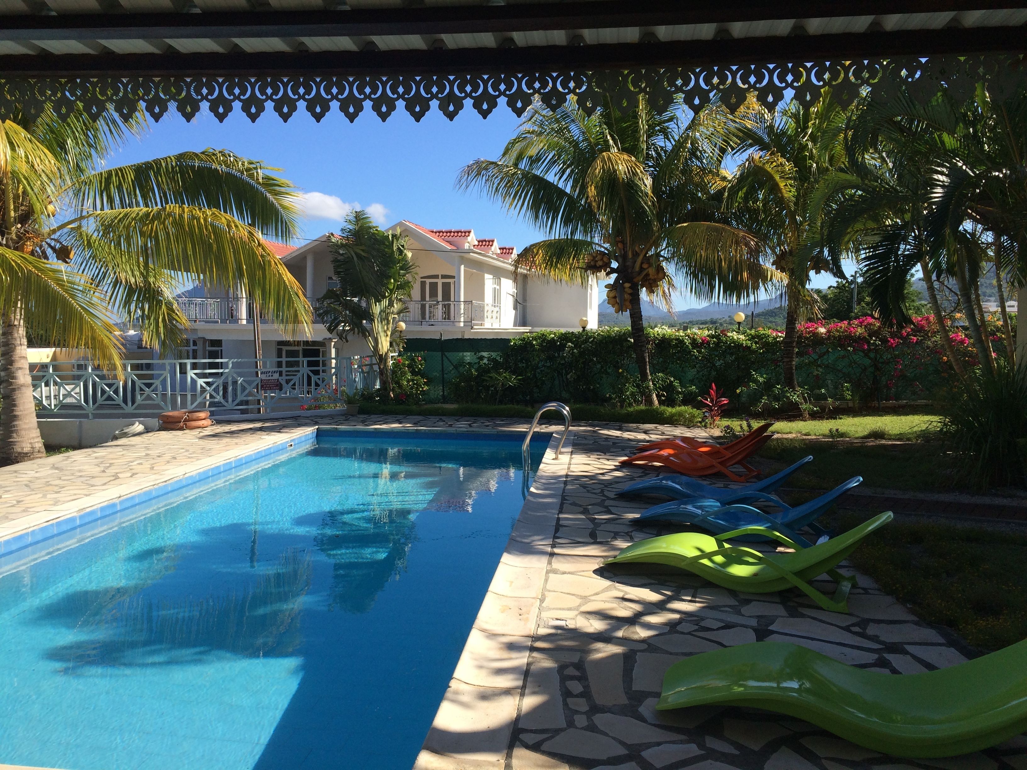 A swimming pool with clear blue water, surrounded by palm trees and a garden with pink flowers.