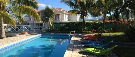 A swimming pool with clear blue water, surrounded by palm trees and a garden with pink flowers.