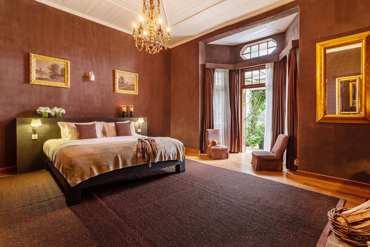 A spacious bedroom with a large bed, a chandelier, and a view of greenery through a window.