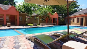 Una piscina con sillas de descanso y sombrillas, rodeada de edificios con tejados rojos y caminos con baldosas.