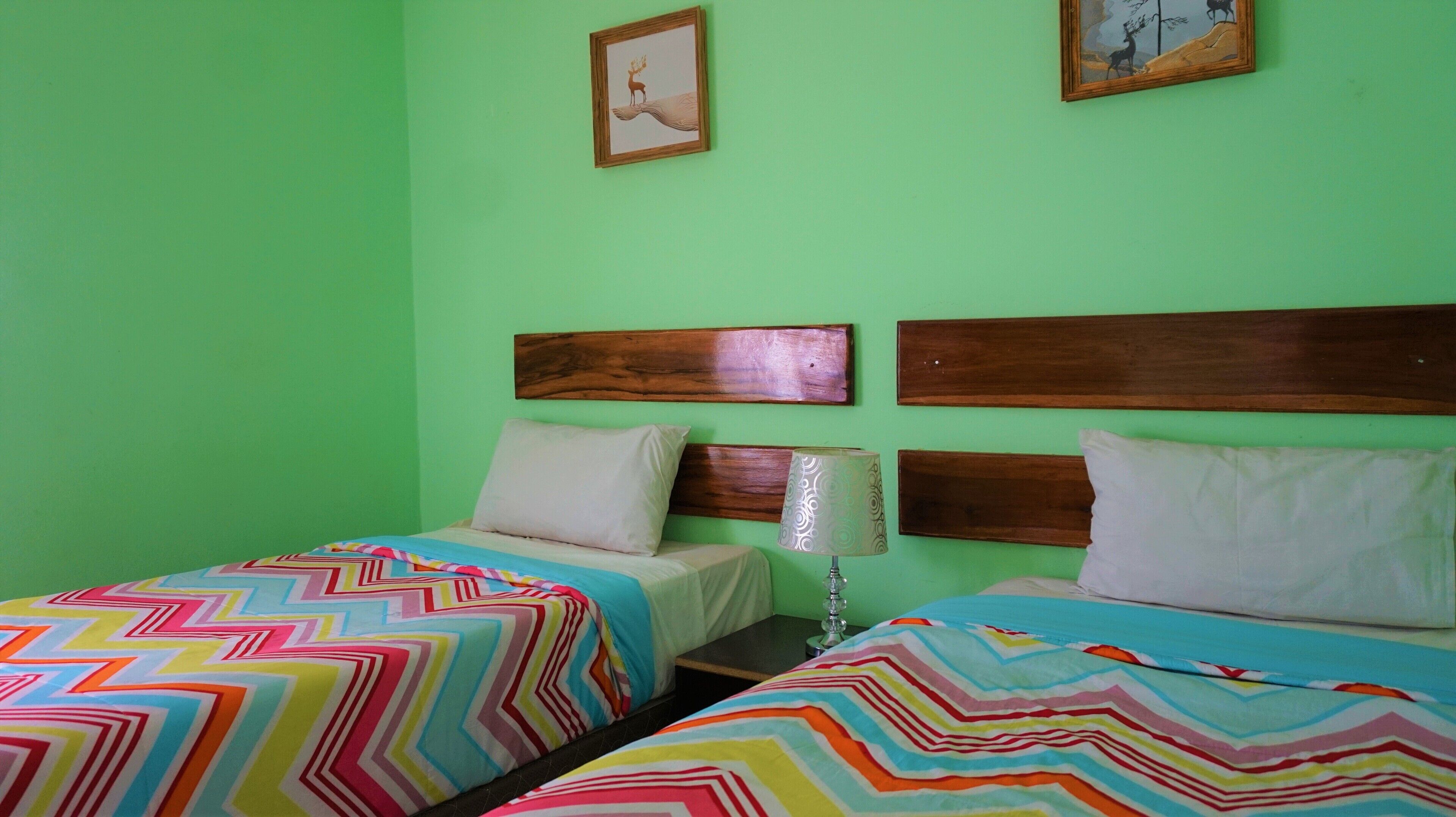 Une chambre avec deux lits, chacun recouvert d’une couvre-lit aux motifs colorés en zigzag. Les lits sont dotés de têtes de lit en bois et une lampe est fixée au mur entre les deux.