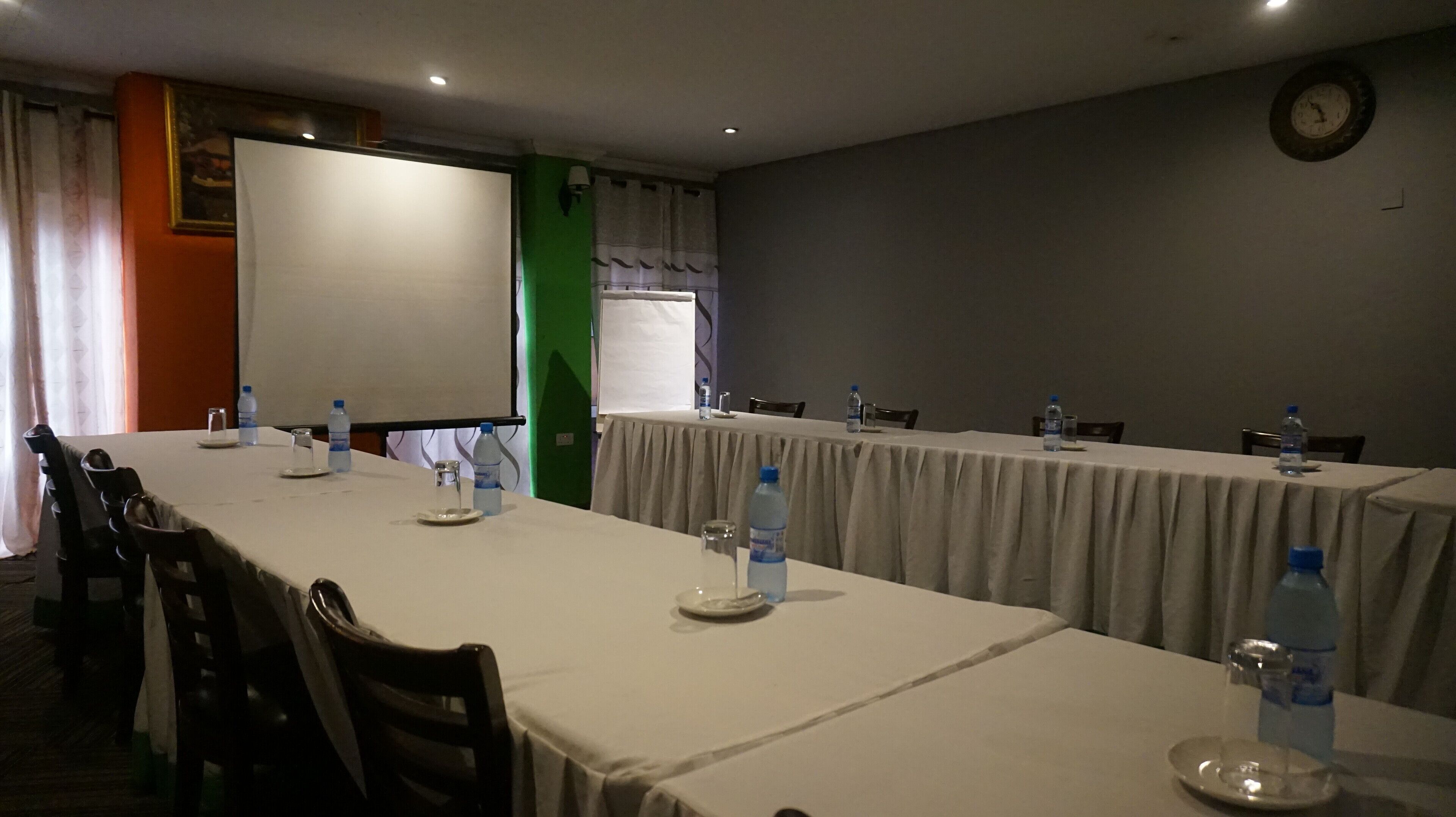 Un salón de conferencias con largas mesas, sillas, botellas de agua y una pantalla de proyector.