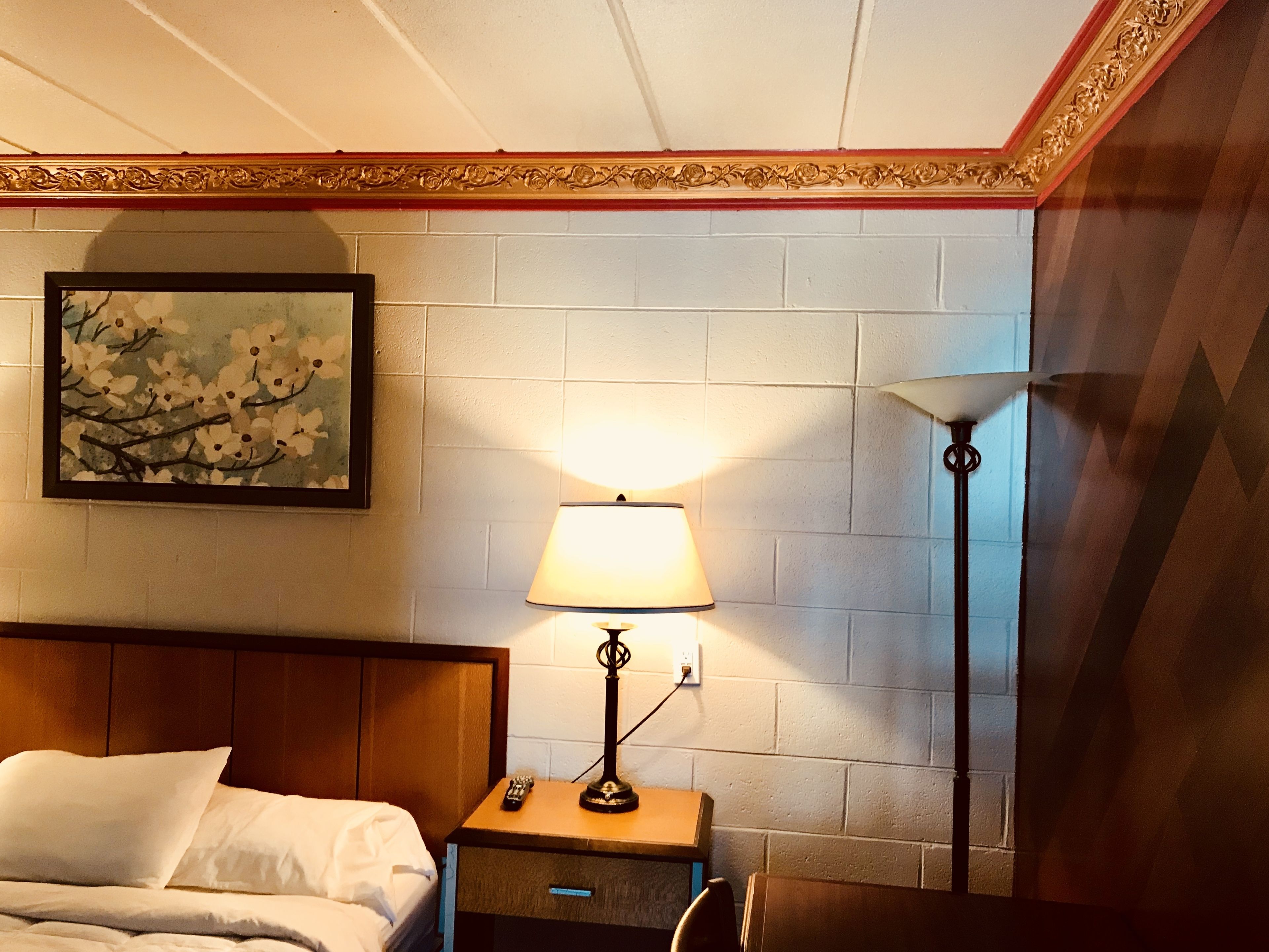 A hotel room with a bed, a nightstand with a lamp, a remote control, and a painting of flowers on the wall.