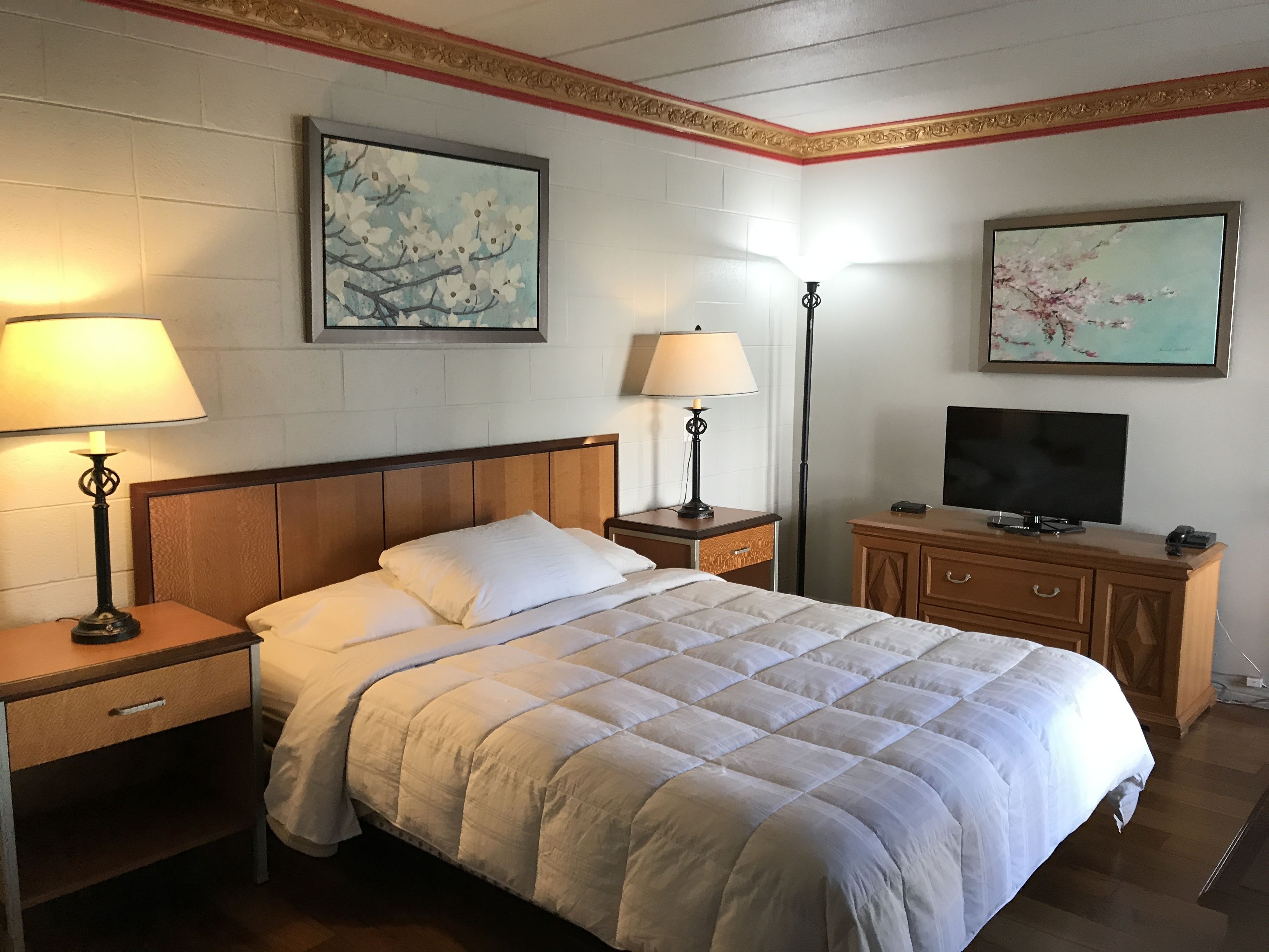 Une chambre d’hôtel comprenant un lit, deux tables de chevet, une commode, une coiffeuse, un téléviseur et deux lampes fixées au mur.