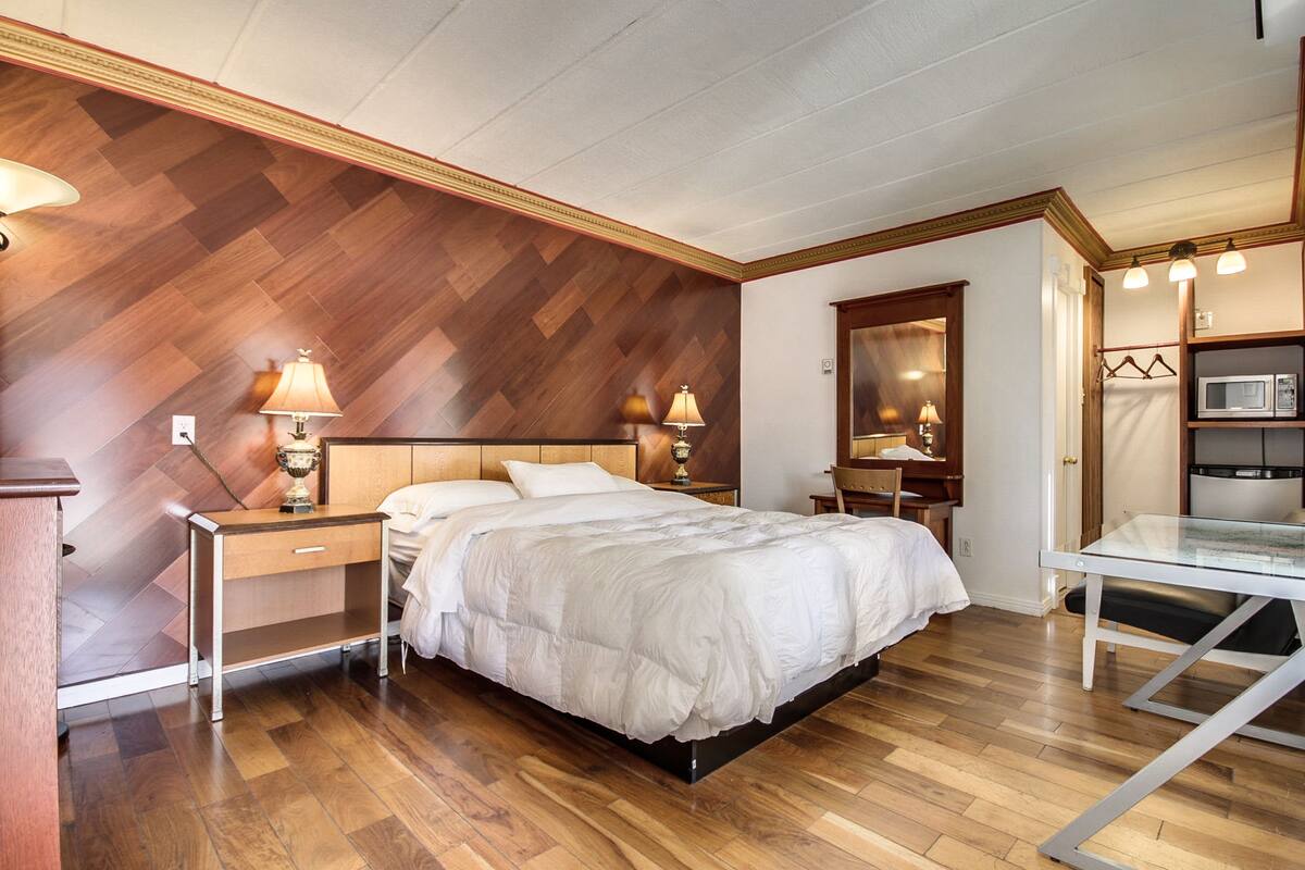 A modern bedroom with a wooden herringbone wall, a bed with white bedding, bedside lamps, a small desk, and a glass table with a chair.