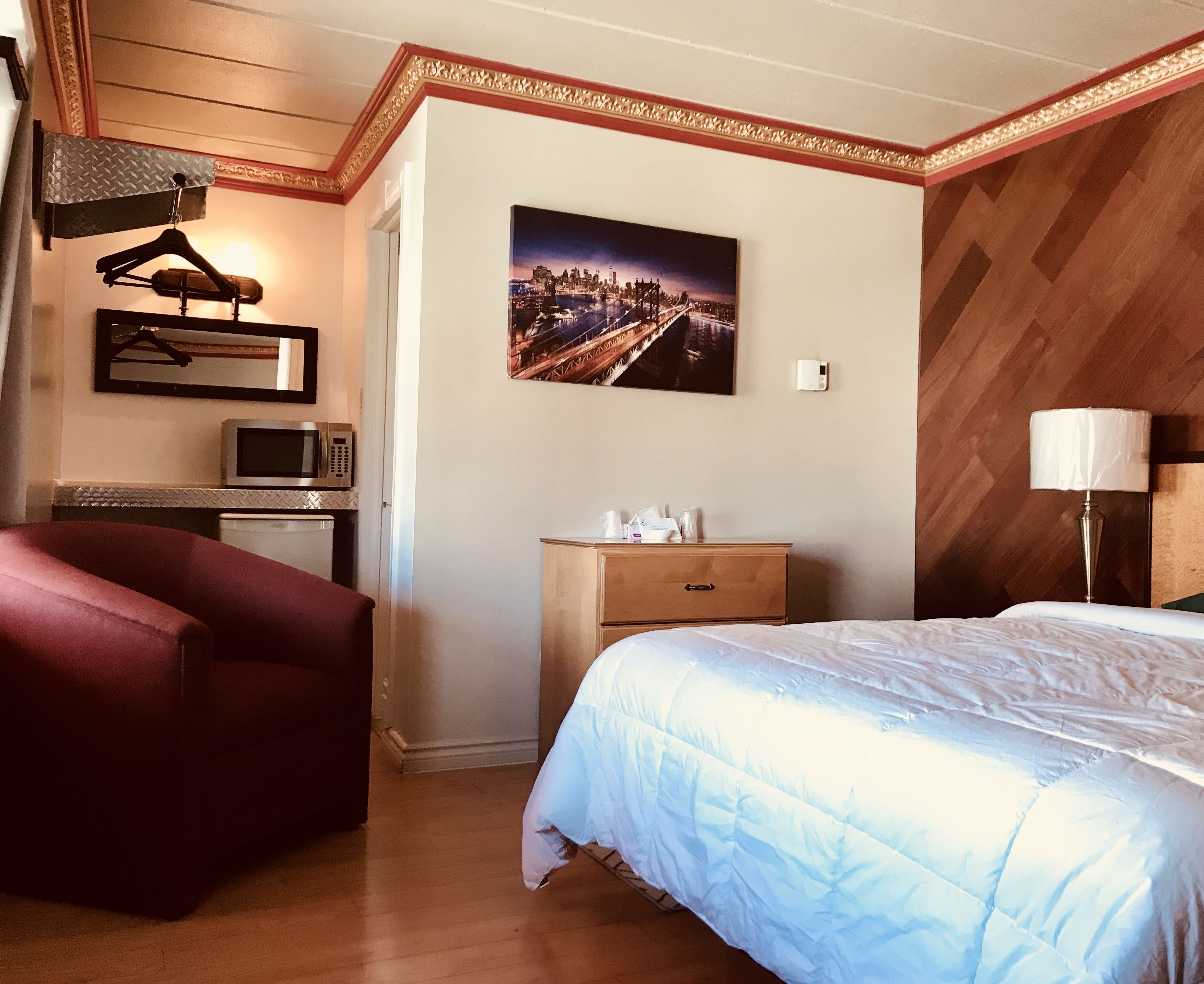 A hotel room with a bed, a desk with a computer, a chair, a dresser, and a painting of a cityscape.