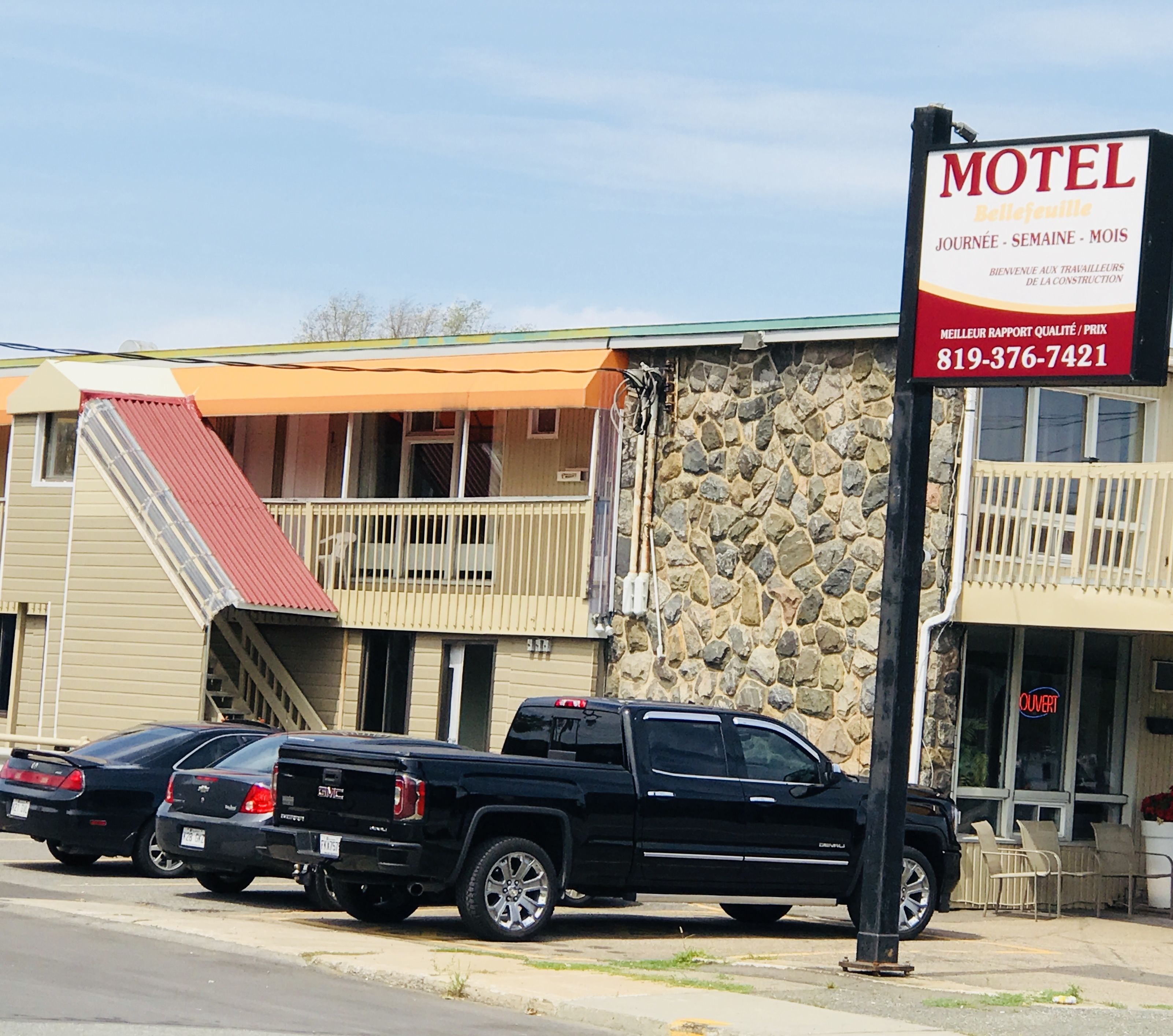 Un motel con fachada de piedra, un toldo rojo y un cartel que lo anuncia como lugar para trabajadores de la construcción. Dos camionetas GMC negras están estacionadas frente a él.