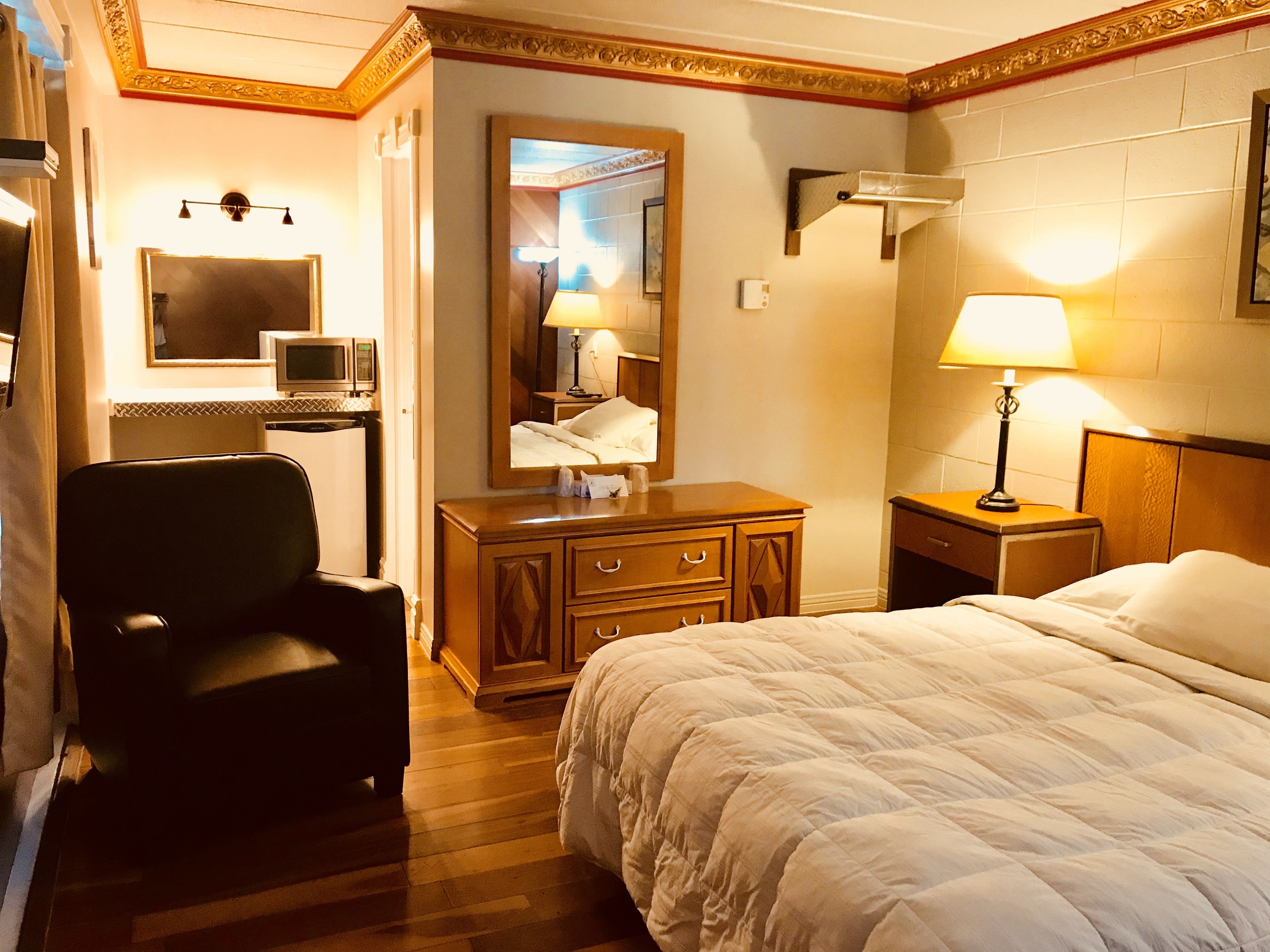 A hotel room with a bed, a desk with a computer, a chair, a dresser, and a nightstand with a lamp.