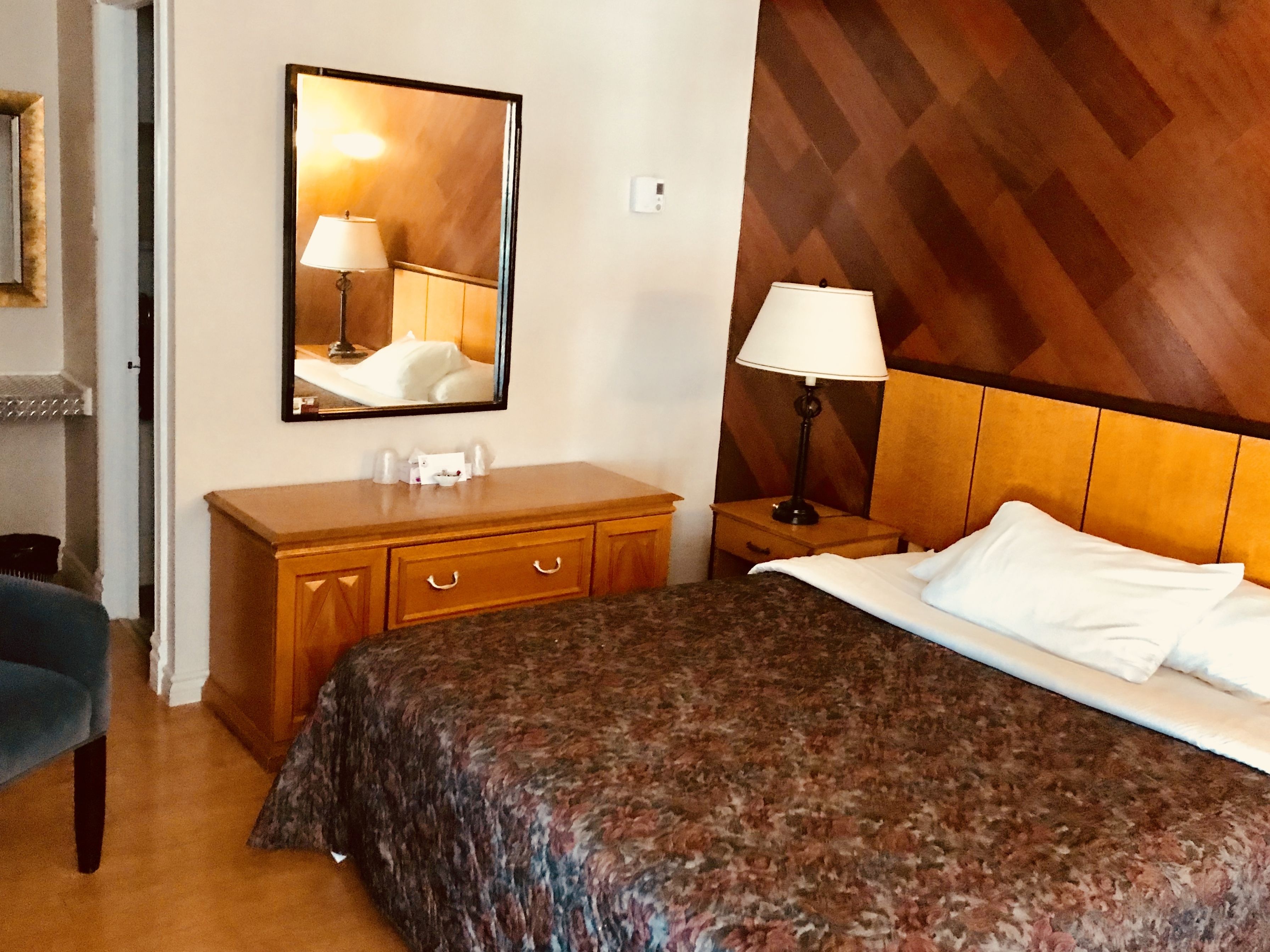 A hotel room with a bed, a wooden headboard, a nightstand with a lamp, a dresser, a mirror, and a chair.