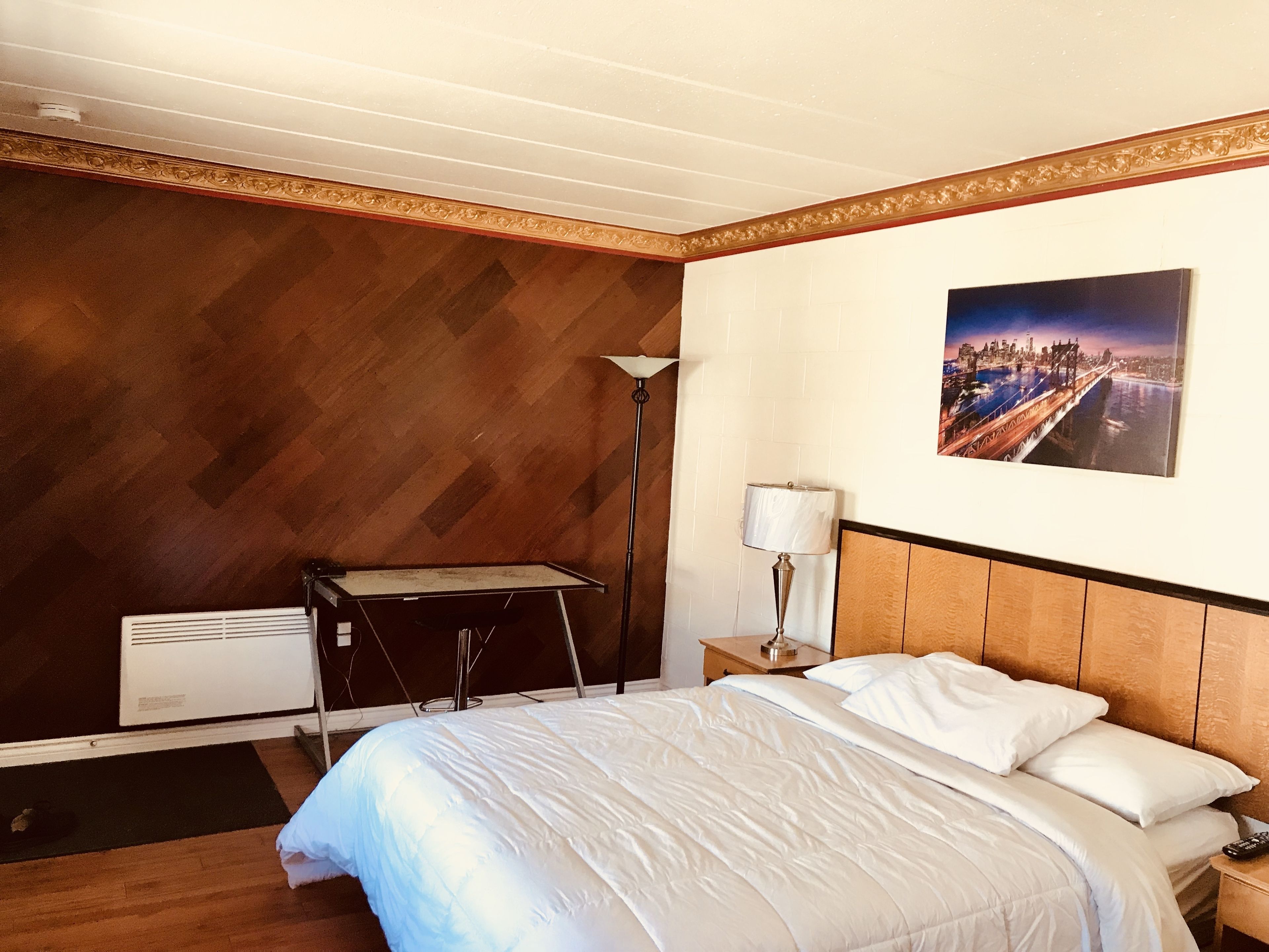 Une chambre d’hôtel avec un lit, une table de chevet, une lampe, un radiateur et un tableau représentant un paysage urbain accroché au mur.