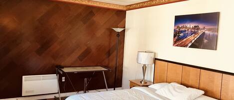 A hotel room with a bed, a nightstand, a lamp, a radiator, and a cityscape painting on the wall.