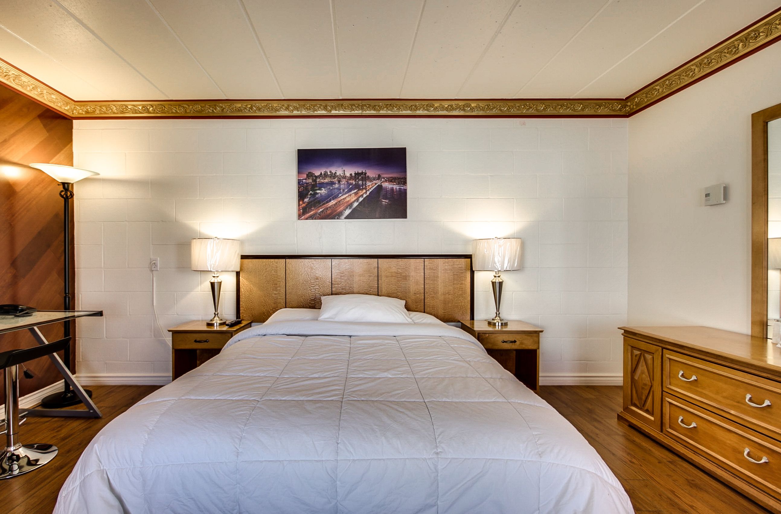 A hotel room with a large bed, two bedside tables with lamps, a wooden dresser, and a cityscape painting on the wall.
