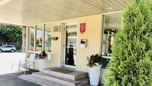 A hotel entrance with a wooden facade, glass doors, and a small outdoor seating area with chairs and potted plants.
