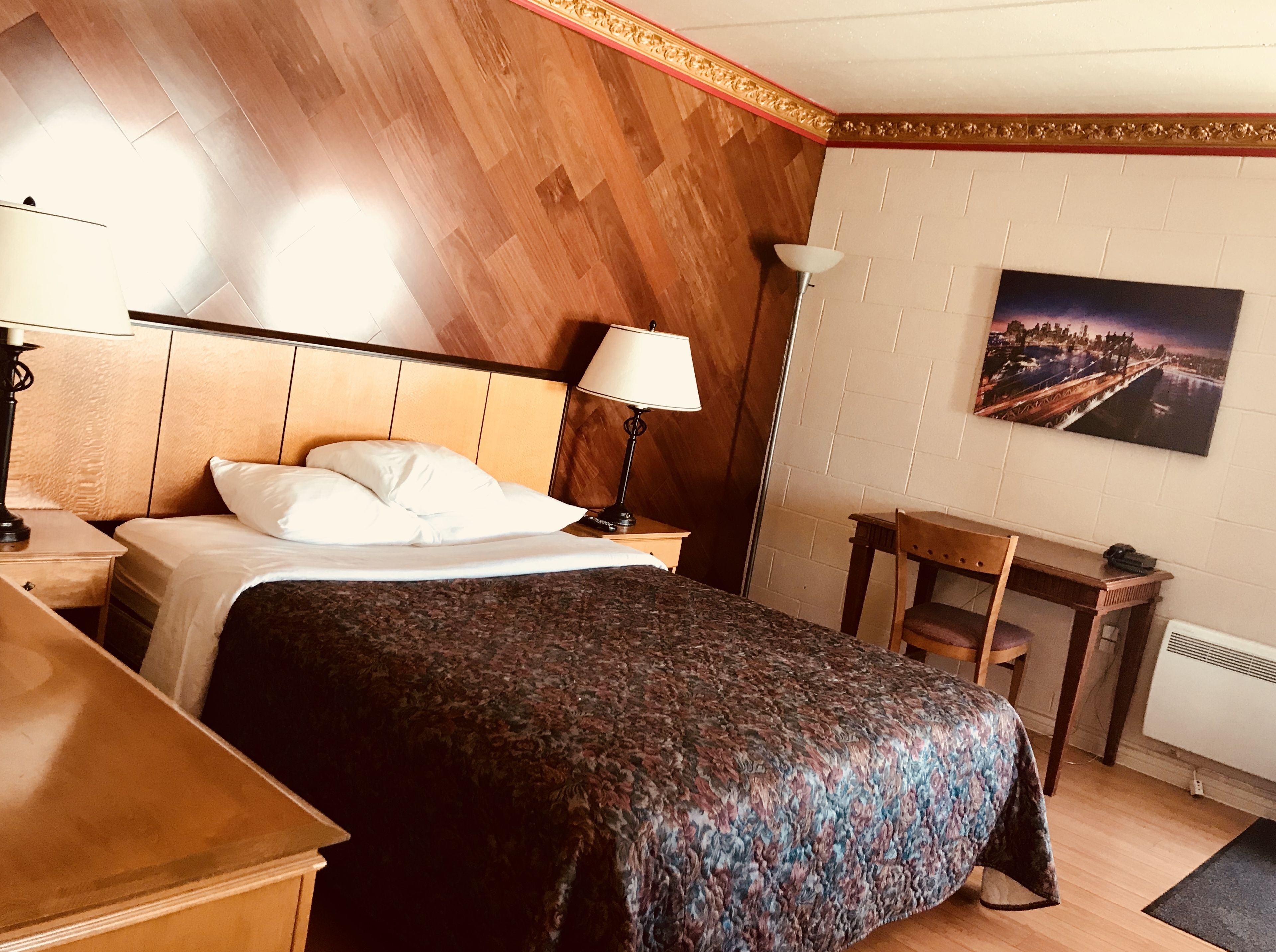 A hotel room with a wooden headboard, a bed with white bedding, a nightstand with a lamp, a small desk with a chair, and a painting on the wall.