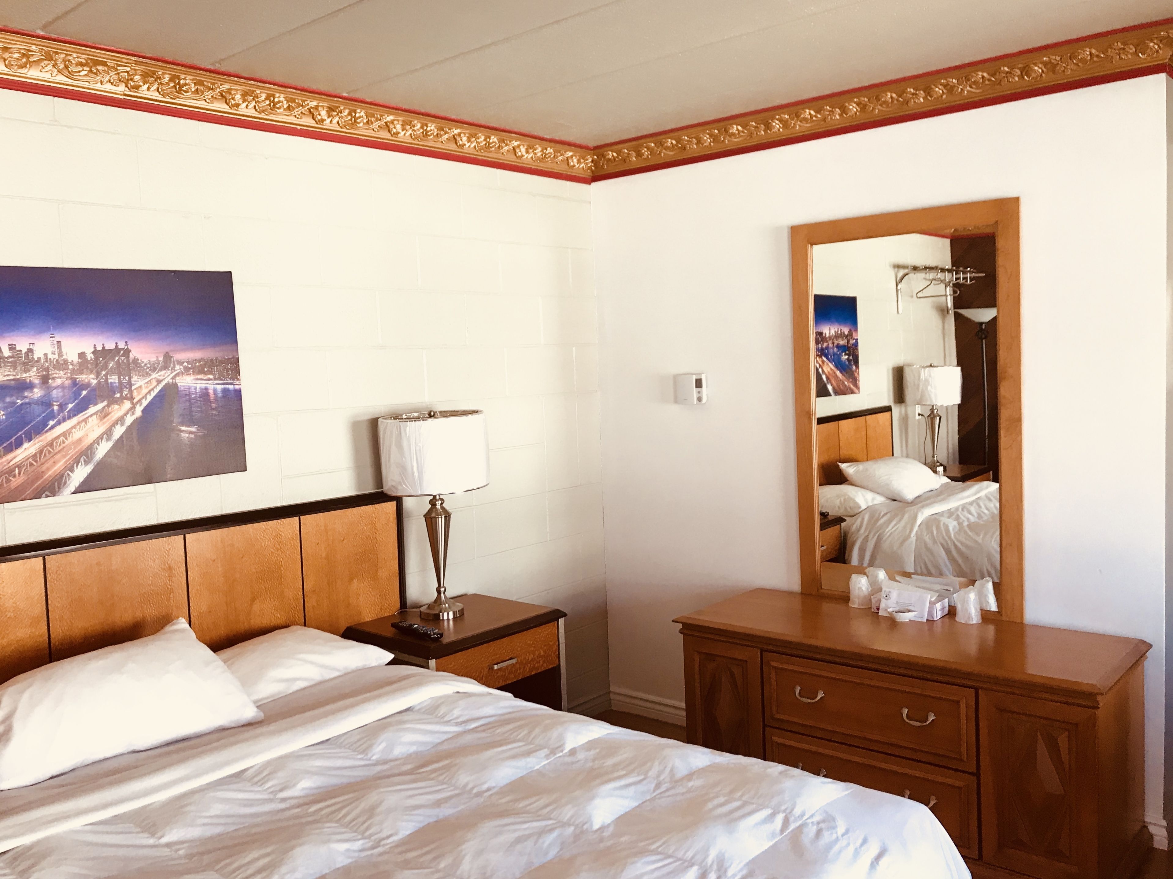 Habitació d'hotel amb un llit, una tauleta de nit, un prestatge, un mirall i un quadre de paisatge urbà a la paret.