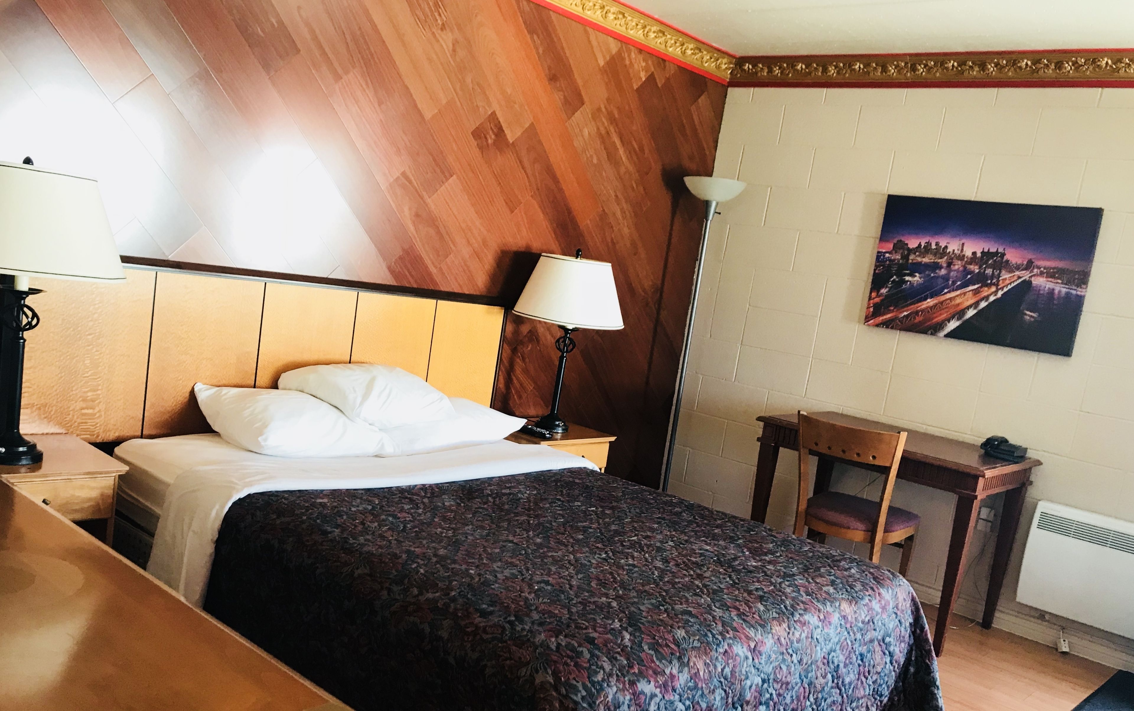 A hotel room with a bed, bedside table, lamp, chair, desk, and a painting on the wall.