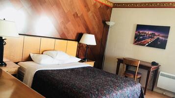 A hotel room with a bed, bedside table, lamp, chair, desk, and a painting on the wall.
