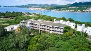 A modern building with a flat roof, surrounded by lush greenery and overlooking a coastal area with a bridge and mountains in the background. - HOTEL E-horizon Resort Condominium Sesoko (Motobu)