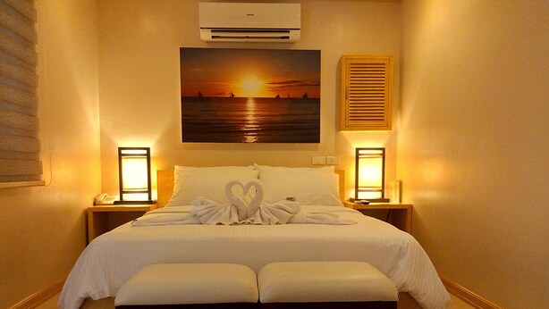 A hotel room with a bed, two bedside tables with lamps, a painting of a sunset, and a window with blinds.