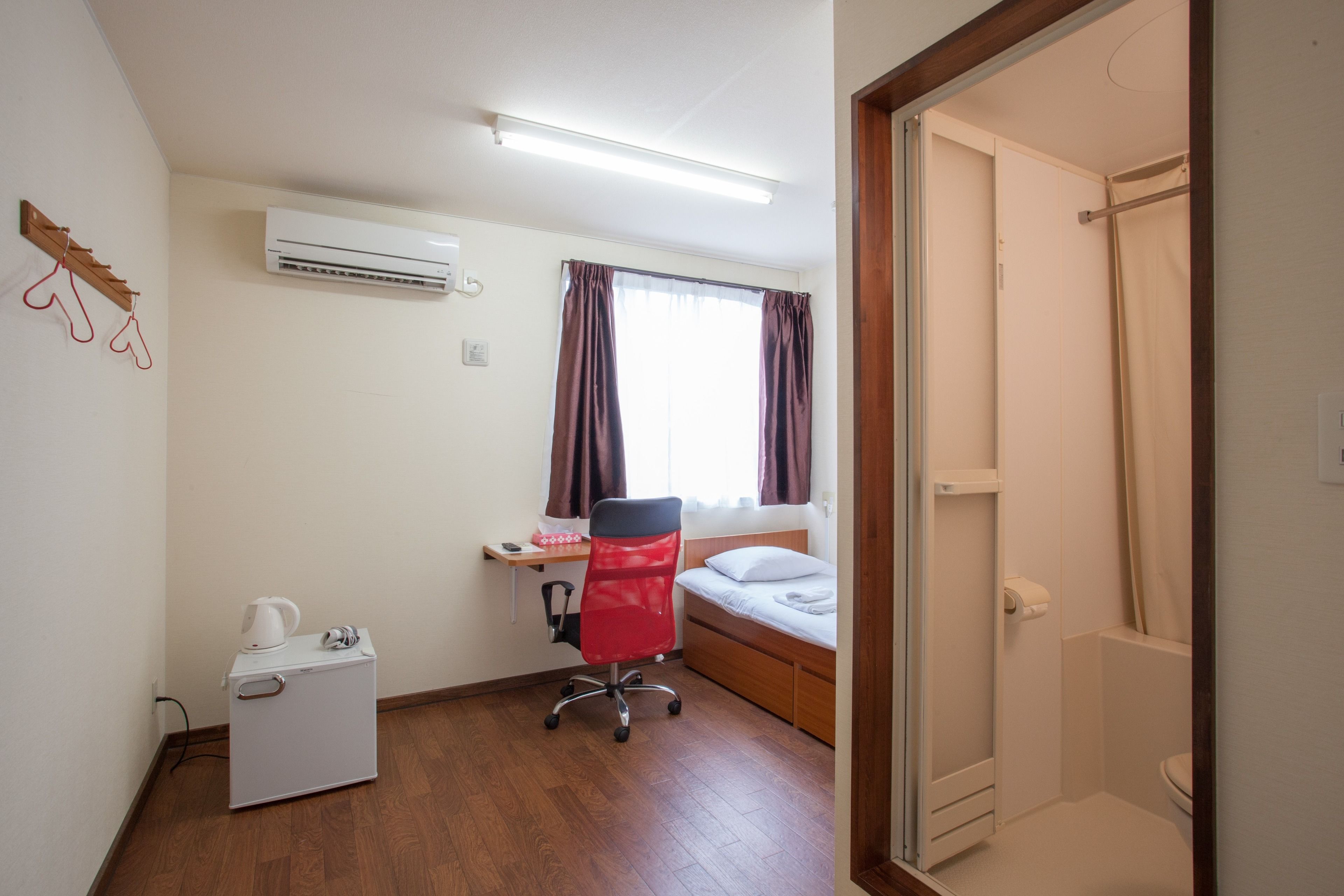 A compact room with a bed, a desk with a chair, a small kitchenette, and a shower area.