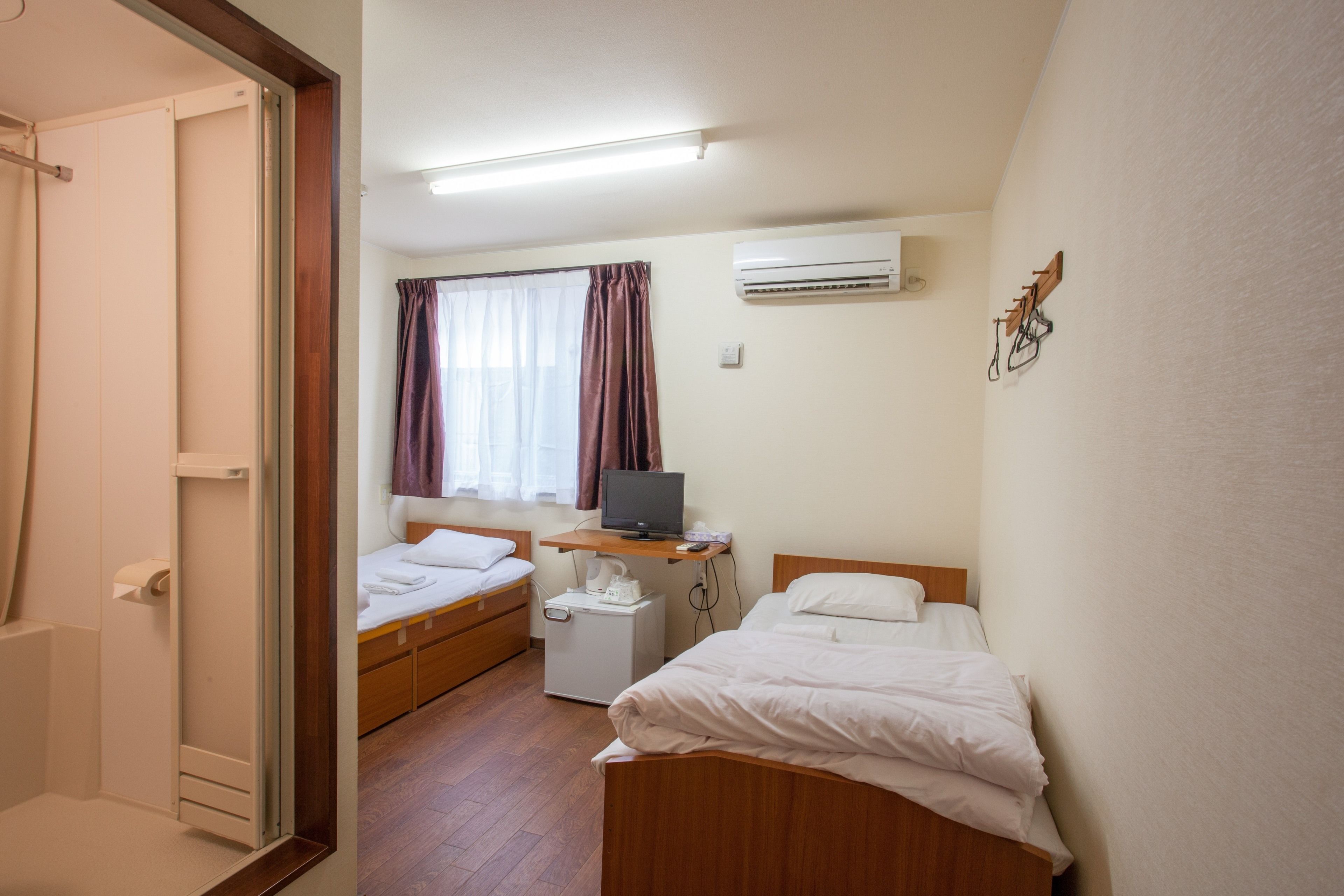 A hotel room with two single beds, a desk with a television, a small white cabinet, and an air conditioning unit on the wall.