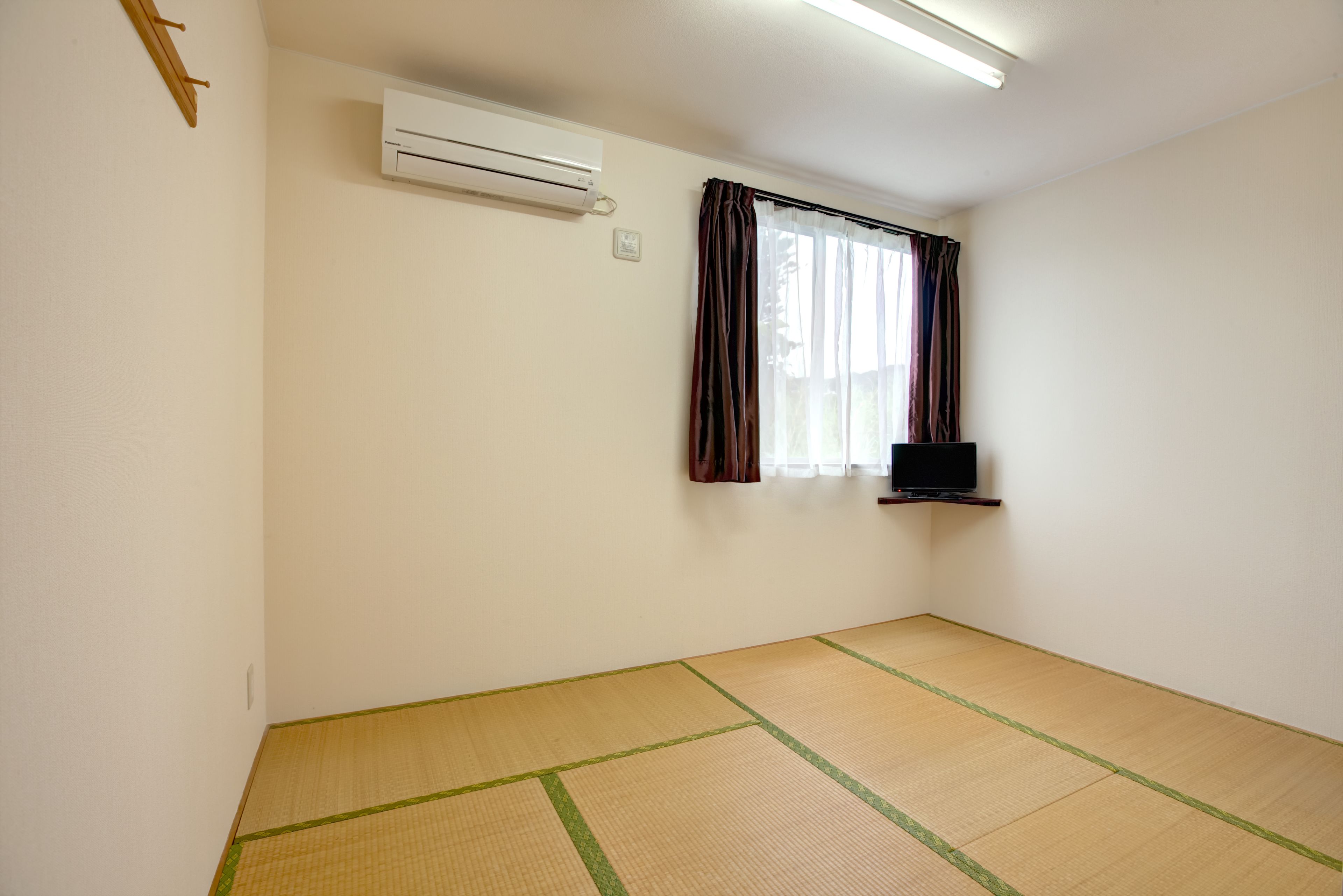 A room with tatami flooring, a mounted air conditioner, a window with curtains, and a flat-screen TV on a shelf.