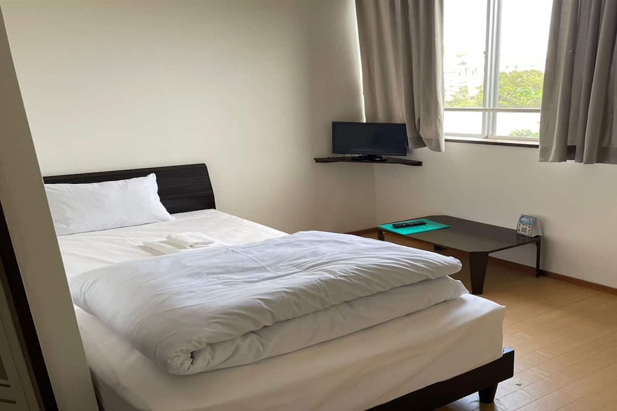 A hotel room with a bed, a TV on a stand, a small table, and a window with curtains.