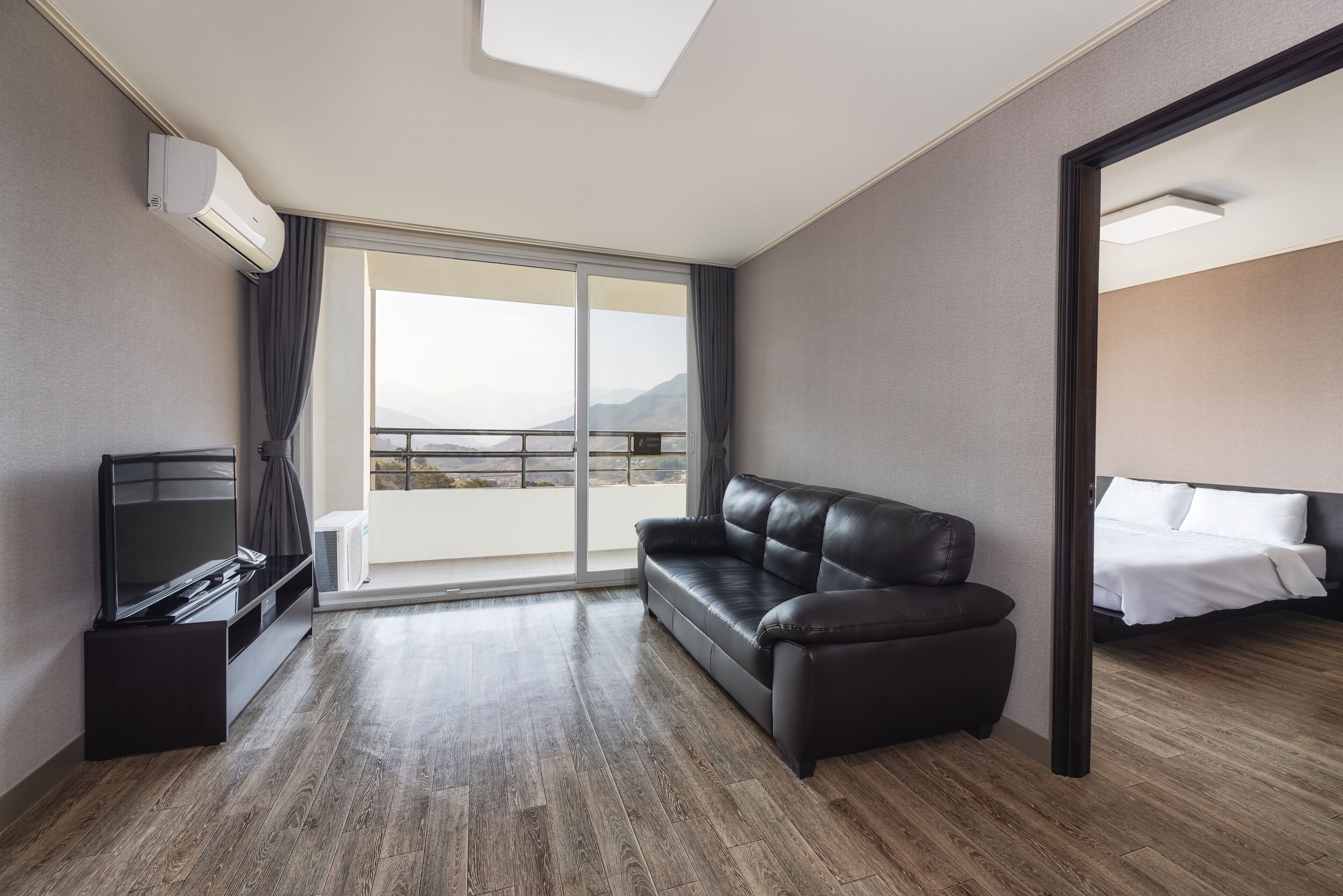 A hotel room with a large sliding glass door to a balcony, a black leather sofa, a flat-screen TV, and a bed with white bedding.