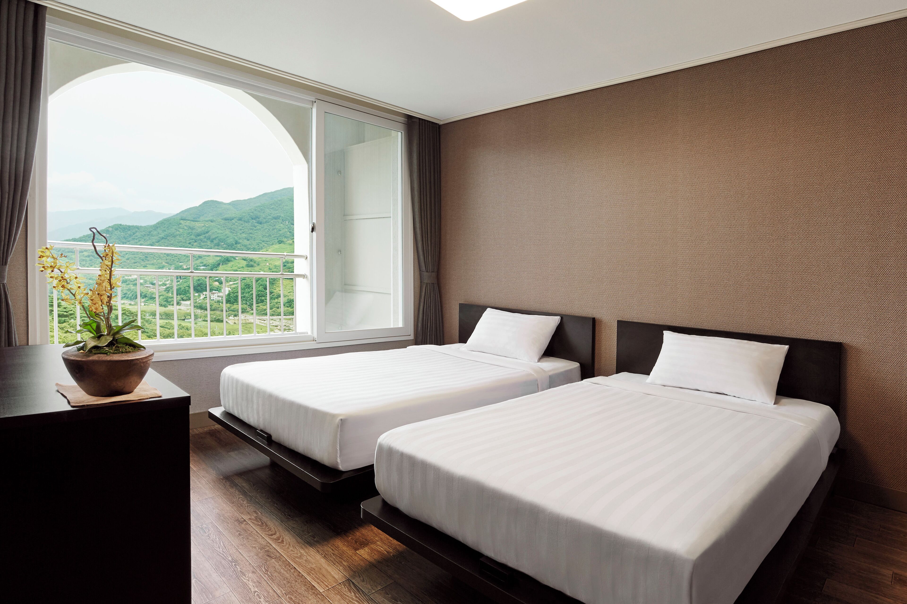 A hotel room with two beds, a desk, and a large window offering a view of green hills.