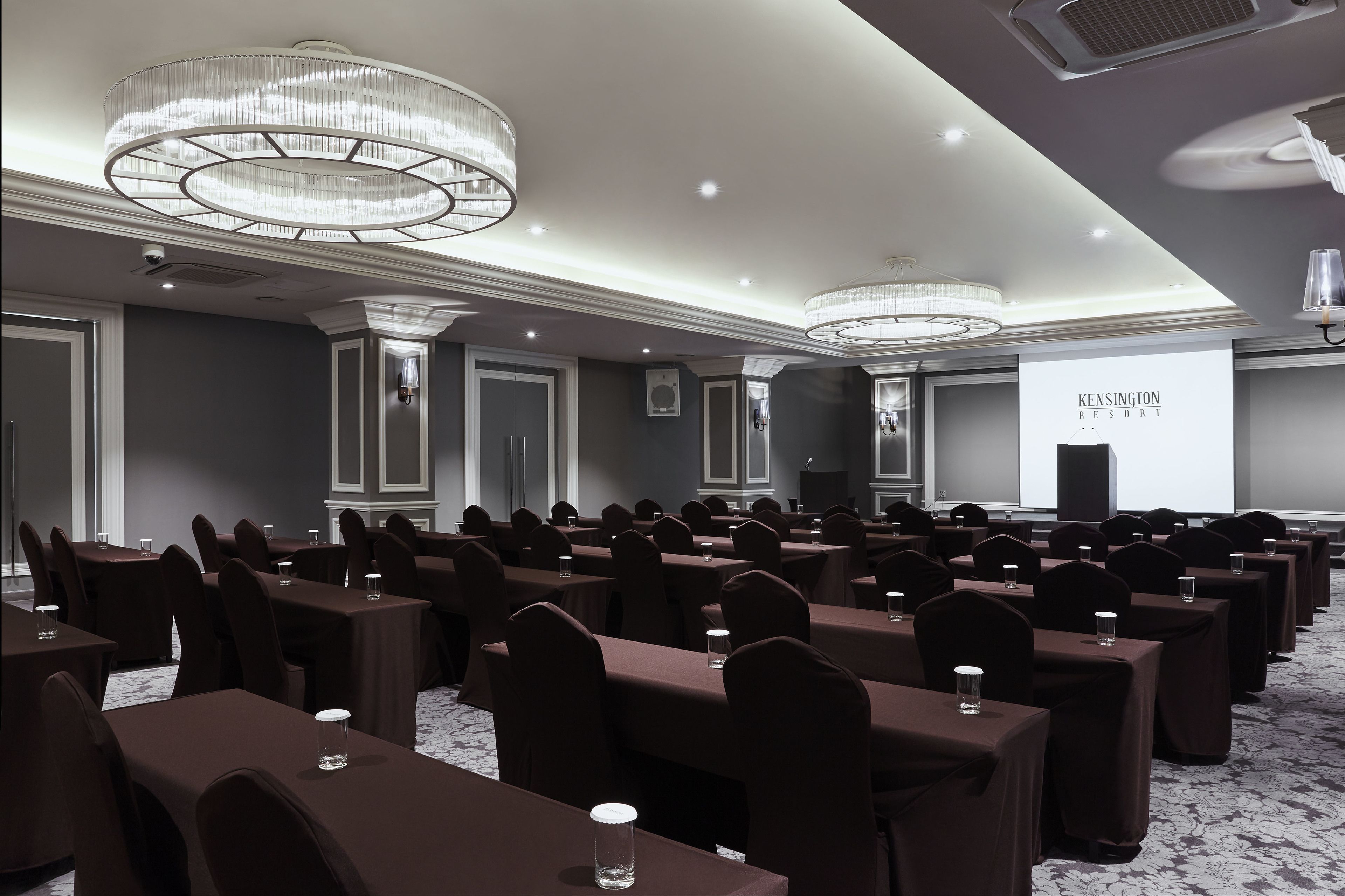 A conference room with rows of tables and chairs, large chandeliers, and a projection screen displaying "Kensington Resort".