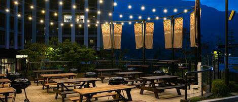 Une terrasse extérieure avec des tables de pique-nique en bois et des guirlandes lumineuses, le tout agrémenté d’une vue sur des montagnes et un bâtiment.