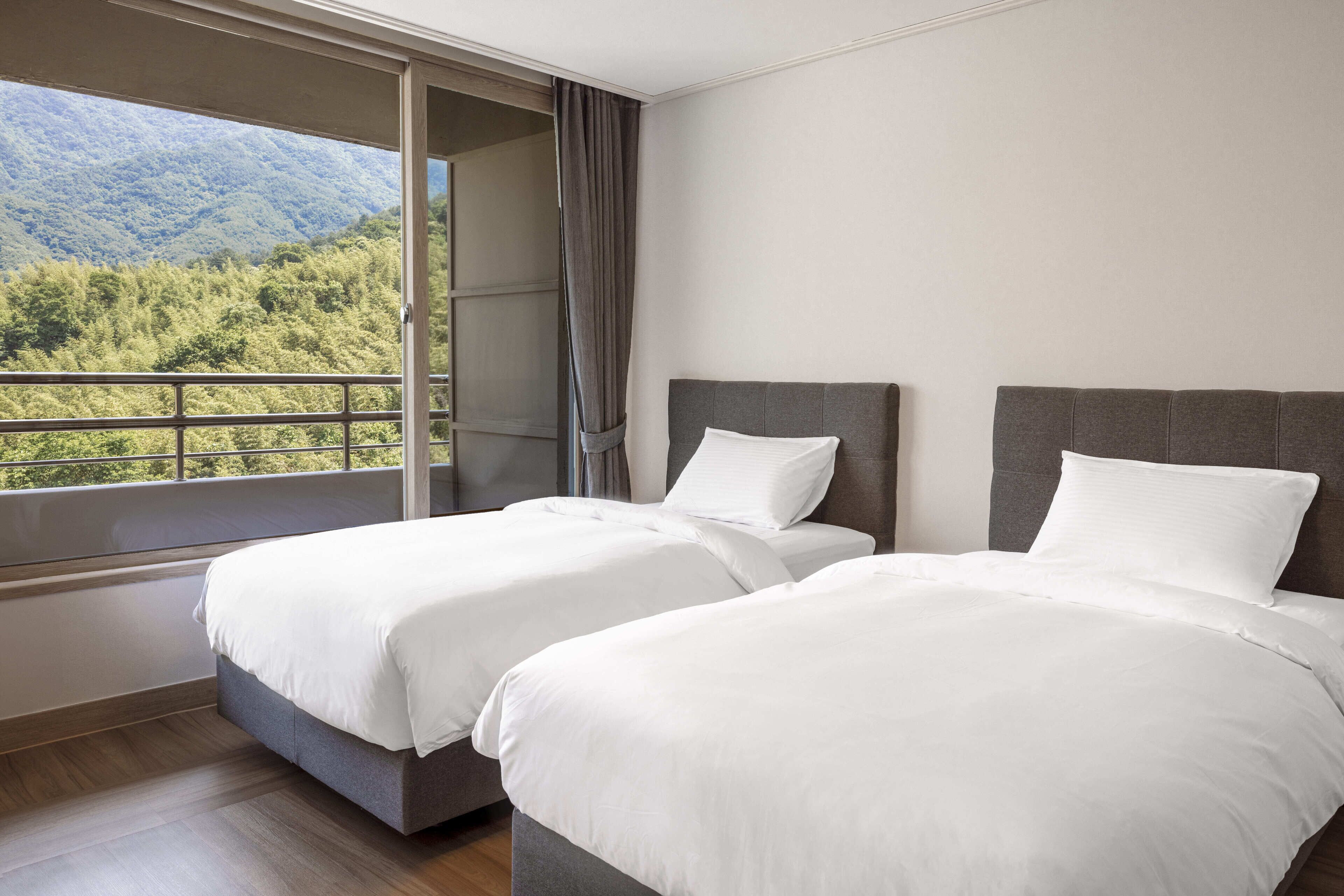 A hotel room with two beds, a large window with a view of a forested mountain, and a balcony with a railing.