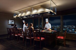 A modern dining area with a chef preparing food at a central island, surrounded by guests seated at wooden tables. - Courtyard by Marriott Shin-Osaka Station (Osaka)
