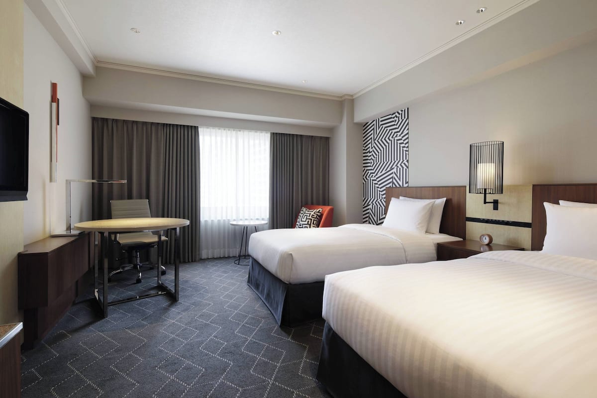 A hotel room with two beds, a desk, a chair, a TV, and a large window with curtains.