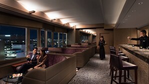 Un espacio lounge moderno con asientos y barra, ventanas amplias con vista a la ciudad, y un barista preparando una bebida.