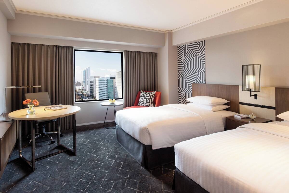 A hotel room with two beds, a desk, a chair, a small table with flowers, and a view of the city.