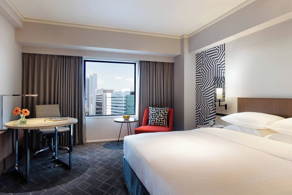 A hotel room with a large bed, a desk with a chair, a small table with flowers, and a view of the city skyline through a large window.