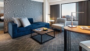 Una habitación de hotel moderna con un sofá azul, una mesita redonda pequeña, una silla y un ventanal grande con vista a la ciudad.