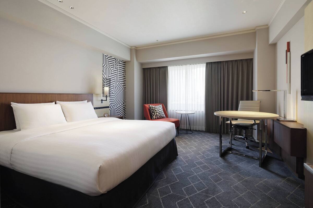 A hotel room with a large bed, a desk with a chair, a small table, and a TV.