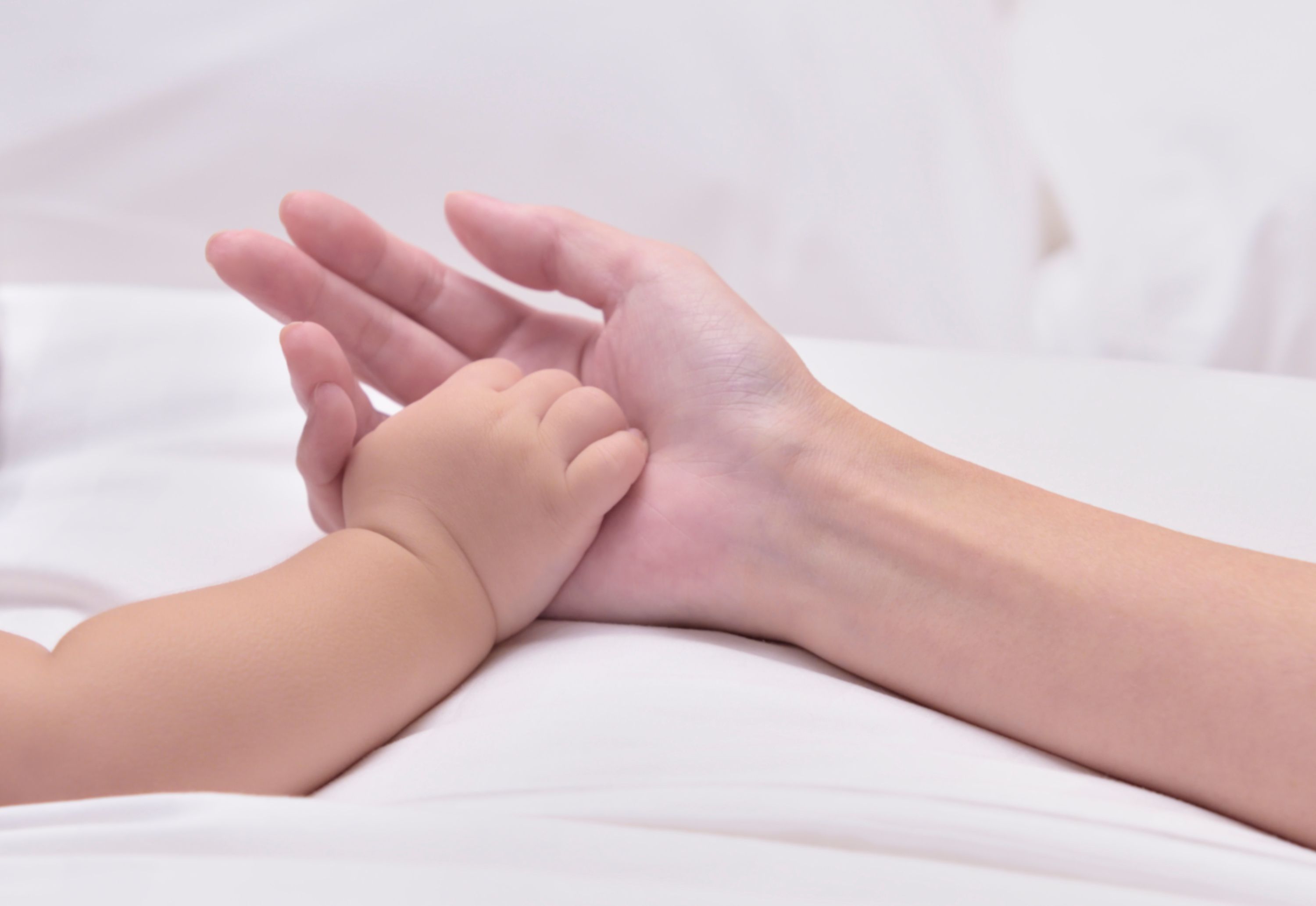 Gros plan d’une main tenant une petite main, probablement celle d’un bébé, sur un lit blanc.
