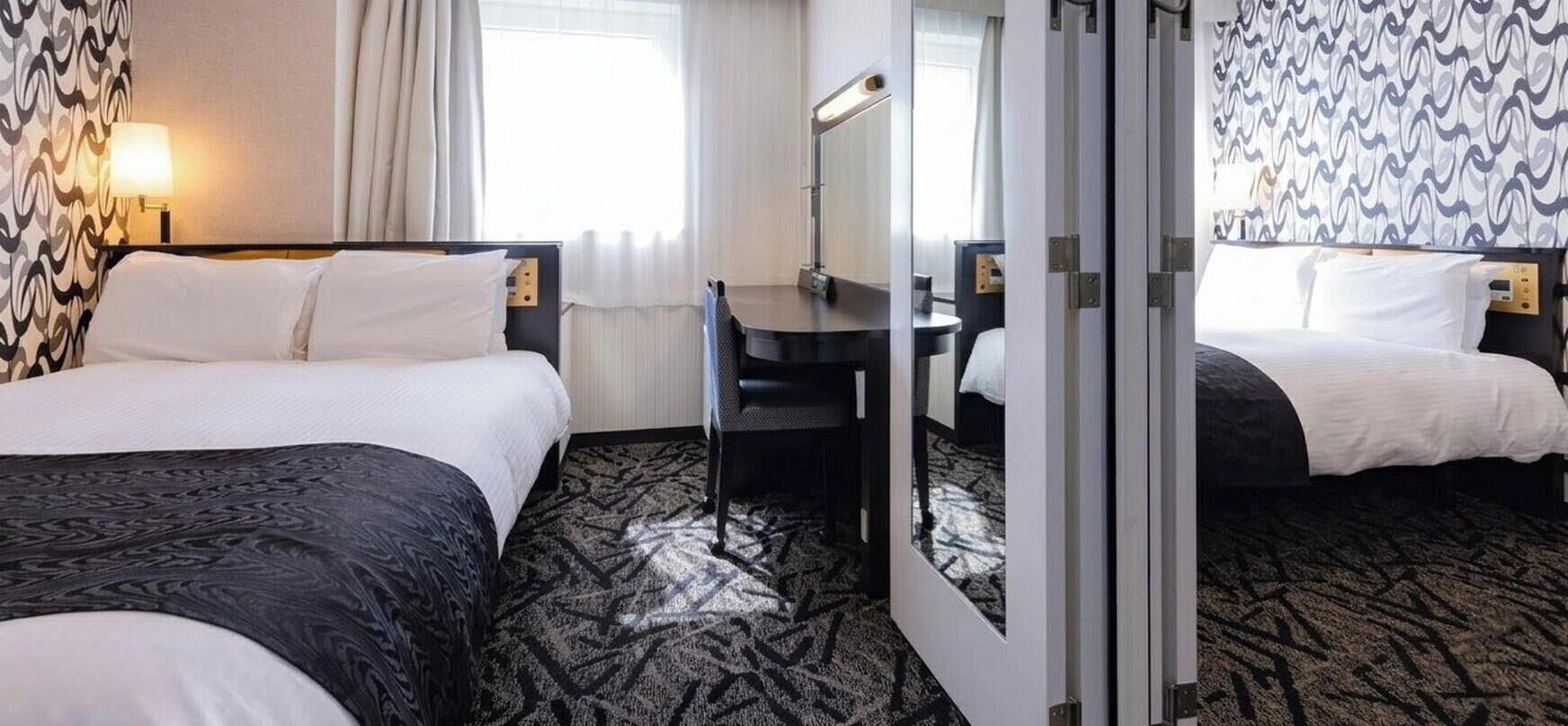 Ein Hotelzimmer mit zwei Betten, einem Schreibtisch, einem Spiegel und einem gemusterten Teppich.